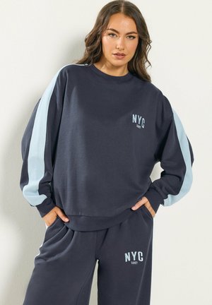 Junge Frau in dunkelblauem Trainingsanzug mit hellblauen Streifen und dem Aufdruck "NYC 1987", steht mit den Händen in den Taschen vor weißem Hintergrund.