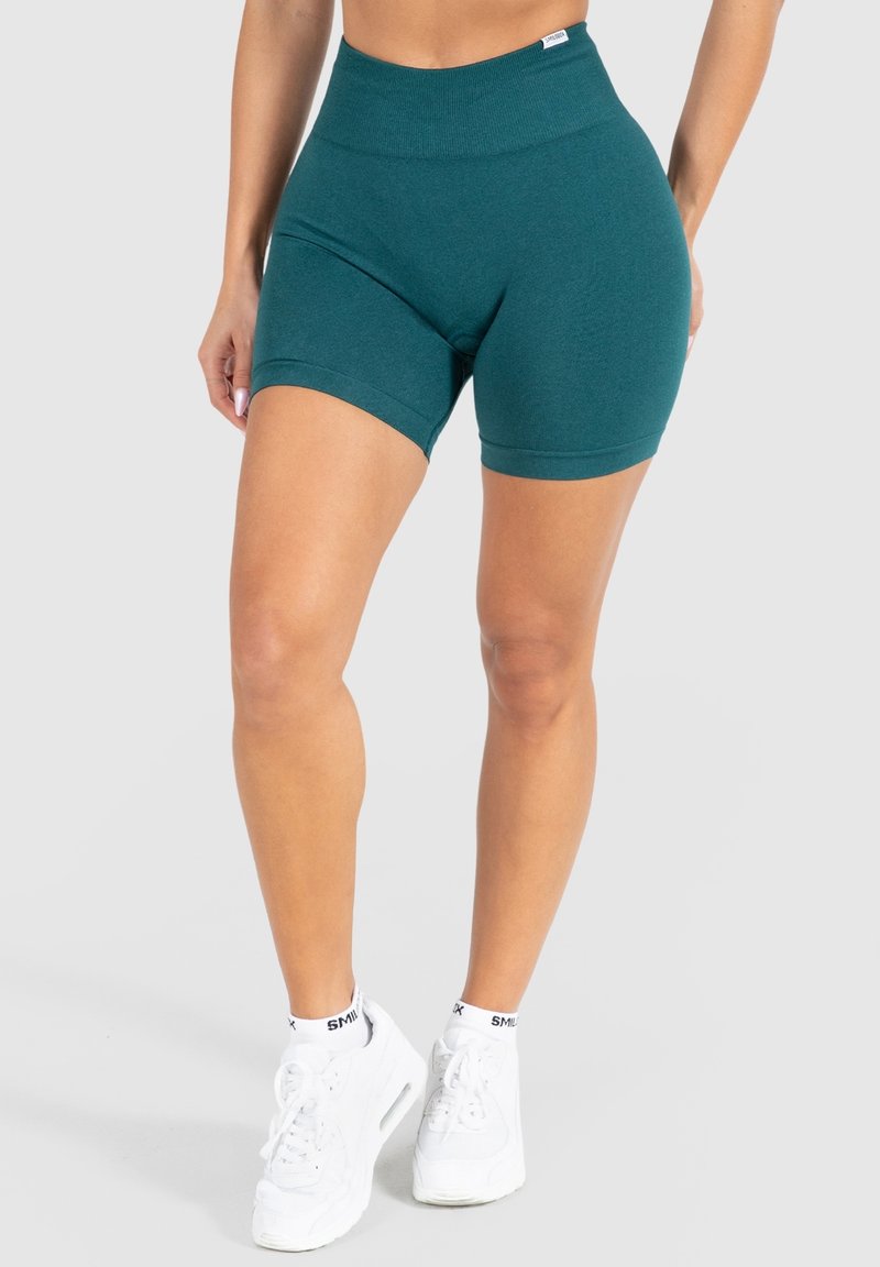 Shorts de sport bleu sarcelle avec une taille haute, une texture lisse et une coupe ajustée. Associés à des baskets blanches et des chaussettes noires retroussées.