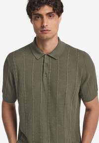 Olivgrön polo med strukturerad stickning och kabelmönster, korta ärmar och klassisk krage. Treknappsslå som detalj.