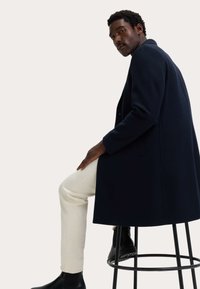 Marineblauwe wollen overjas, getailleerde pasvorm, gedragen over beige broek. Zwarte Chelsea boots. De persoon zit op een zwarte kruk tegen een neutrale achtergrond.