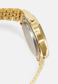 Montre dorée avec un bracelet en maillons texturés, forme de boîtier octagonal et dos rond en argent. Présente un bouton latéral pour les réglages.