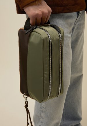 Mano sosteniendo una pequeña bolsa rectangular verde y marrón con cremalleras dobles plateadas y un cordón en bucle adjunto, sobre un fondo de vaqueros de lavado claro.