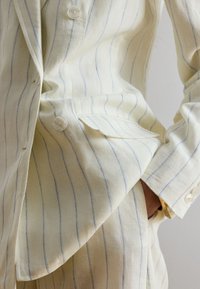 Inimene, kes kannab kreemivärvi ülikonda, millel on õhukesed sinised vertikaalsed triibud, käsi taskus ja kuldne käevõru randmel, lähivaade.