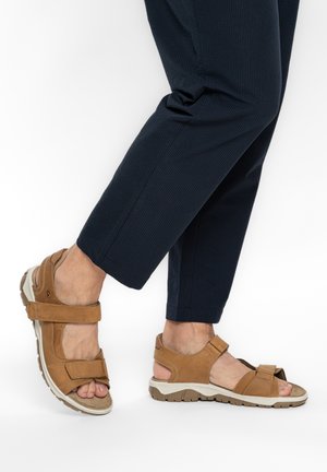 Füße in braunen Sandalen mit verstellbaren Riemen, kombiniert mit dunkelblauen Hosen, ein Fuß leicht angehoben vor weißem Hintergrund.