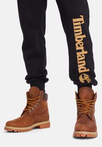 Stivali in pelle marrone con punta rotonda, collo imbottito, lacci arancioni e suola in gomma. Pantaloni sportivi neri con logo "Timberland" in oro.