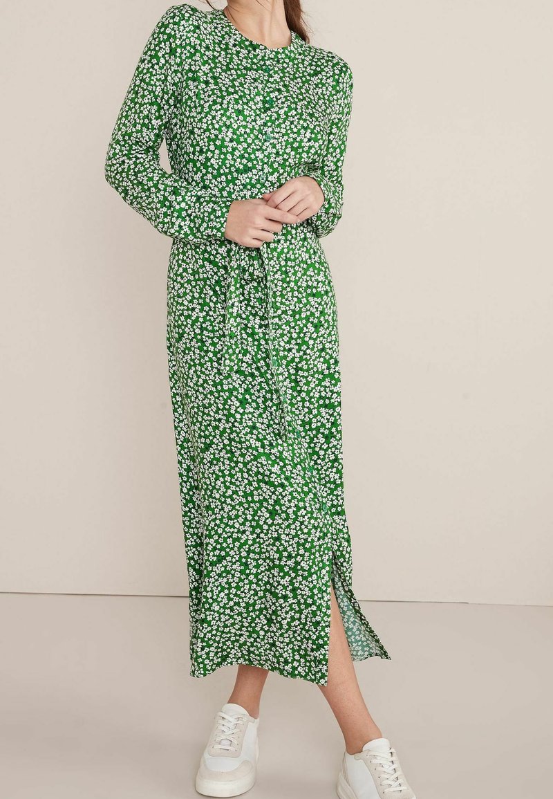 Robe maxi à imprimé floral vert avec manches longues, encolure ronde, lien à la taille et fente sur le côté. Associée à des baskets blanches.