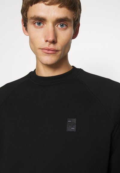 Sweatshirt noir avec col en côte et manches raglan. Patch logo en relief remarquable sur la poitrine. Texture de tissu lisse, design minimaliste.