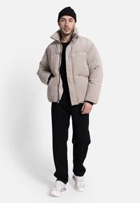 Beige corduroy puffer jacket with large pockets, high collar, and a textured finish, paired with black pants and white sneakers.