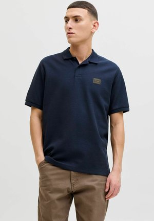 Uomo che indossa una polo a maniche corte blu scuro con un piccolo logo frontale, pantaloni marroni, mani in tasca, che guarda di lato.