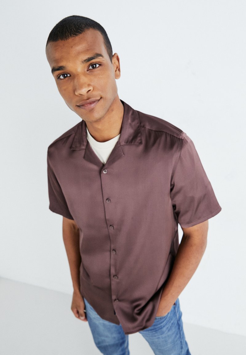 Chemise en satin marron à manches courtes avec un devant à boutons, texture douce et col décontracté, portée par-dessus un t-shirt de couleur claire.