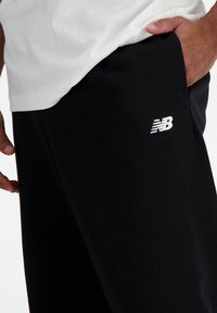 Schwarze Jogginghose aus einem weichen Stoff, mit einem weißen "NB"-Logo auf dem Oberschenkel und seitlichen Taschen für Funktionalität.