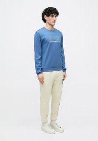 Zils džemperis ar apaļu izgriezumu un grafisku logotipu, kombinēts ar krēmkrāsas sporta biksēm. Modelis valkā baltas sporta kurpes. Vienkāršs, ikdienišķs dizains.