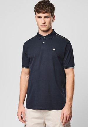 Młody mężczyzna w granatowej koszulce polo z białym wykończeniem na rękawach i ramionach, stojący na tle jednolitego jasnoszarego tła.