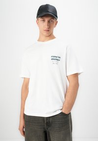 Ung mann iført en hvit grafisk t-skjorte med tekst, svart caps og mørke jeans, stående mot en helt hvit bakgrunn.