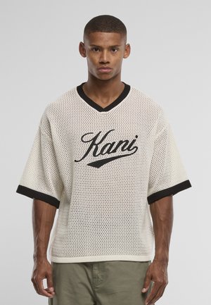 Jeune homme portant une chemise en mesh blanche oversize avec le texte "Kani" en noir et une bordure noire aux manches et au col, se tenant devant un fond uni.