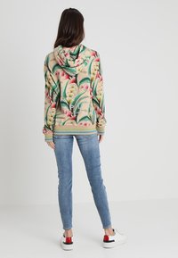 Huvtröja med blommönster i rosa och grönt på en beige bakgrund. Ribbad mudd och fåll, bärs med ljusblå jeans och sneakers.