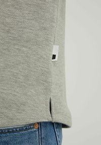 Graues Baumwoll-T-Shirt mit abgerundetem Saum, seitlichen Schlitzen und einem kleinen schwarz-weißen Etikett an der Unterseite. Getragen mit blauer Jeans.