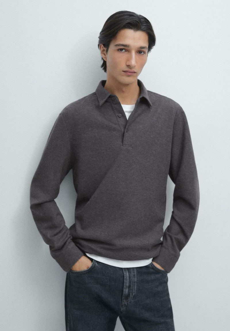 Donkergrijze trui met een kraag en een drieknoopsbies, gedragen over een wit overhemd, gecombineerd met donkere denim jeans. Zachte, textuurrijke stof.