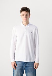 Langermet hvit polo skjorte laget av bomull, med klassisk krage, tre knapper og en liten svart logo på venstre bryst.