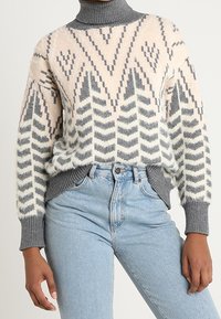 Stickad tröja med hög krage, geometriska mönster i kräm, grått och rosa. Ärmarna är ribbade, vilket ger textur och struktur.