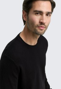 Mann mit kurzen dunklen Haaren und Bart, der einen schwarzen Rundhalsausschnitt-Pullover trägt und leicht zur Seite schaut vor einem schlichten hellen Hintergrund.