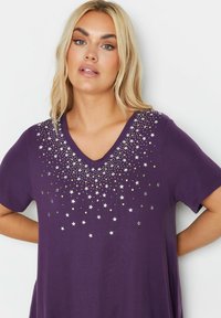 Violetti v-kaula-aukollinen t-paita lyhyillä hihoilla, jossa on hopean ja kullan värisiä tähtimuotoisia koristeita, jotka luovat gradienttimaisen kuvion kaula-aukolla.