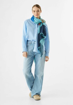Chemise bleu clair à boutons, jean bleu clair à coupe décontractée, chaussures beige à enfiler ; portant une écharpe à motifs multicolores avec des bords frangés.