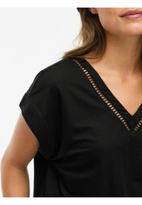 Frau trägt ein schwarzes V-Ausschnitt-Oberteil mit dekorativem Spitzebesatz am Ausschnitt und kurzen umgeschlagenen Ärmeln vor einem schlichten Hintergrund.