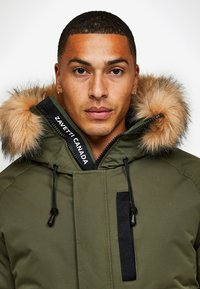 Zaļā parka ar kažokādu oderi kapucē, ar rāvējslēdzēju, melniem akcentiem un logotipa birku. Tekstūrēta auduma ar auklām pie apkakles un apakšmalas.