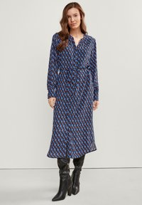 Frau trägt ein gemustertes blau-schwarzes Midikleid mit langen Ärmeln und Taillengurt, kombiniert mit schwarzen, kniehohen Stiefeletten mit Absatz, stehend vor einem einfarbigen Hintergrund.