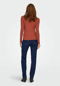 Langärmeliges, geripptes Oberteil in rostrot, kombiniert mit dunkelblauen Jeans. Die Jeans haben einen klassischen Schnitt und minimale Nahtdetails auf der Rückseite.