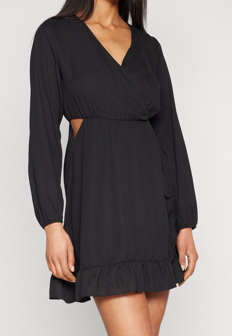 Femme portant une robe noire à manches longues avec un col en V, une taille élastique, des découpes latérales et un ourlet volanté.