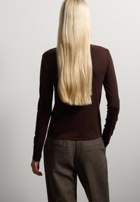 Bruine lange mouwen top gemaakt van zacht materiaal, gecombineerd met grijze gestructureerde broek. Haar valt over de schouders en wordt omlijst tegen een effen achtergrond.