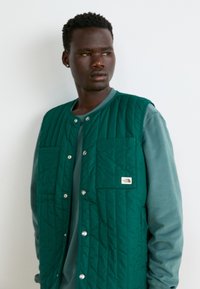 Quilted green vest with two front pockets and silver snap buttons, layered over a gray long-sleeve shirt. Brand logo on the pocket.