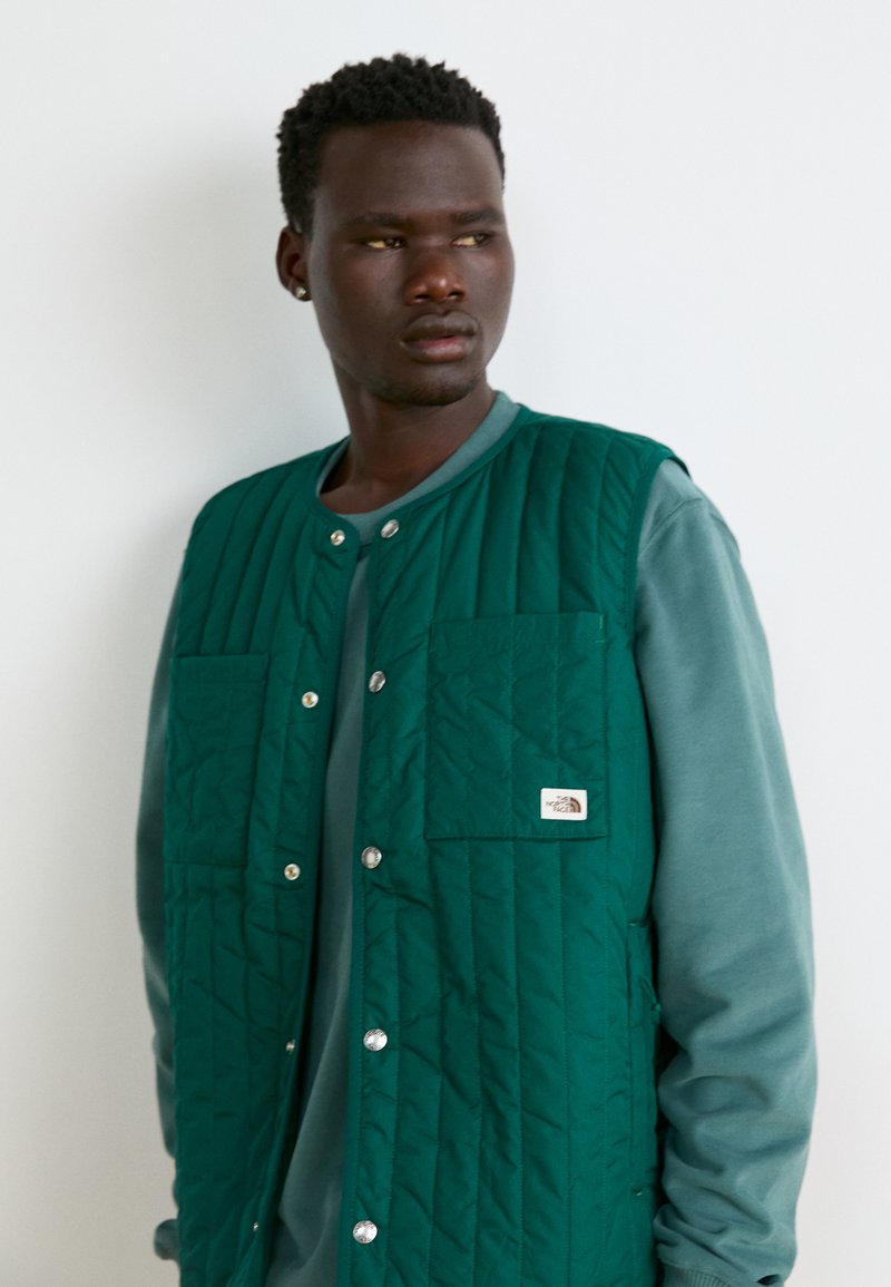 Prošívaná zelená vesta s dvěma předními kapsami a stříbrnými druky, vrstvená přes šedou košili s dlouhým rukávem. Logo značky na kapse.