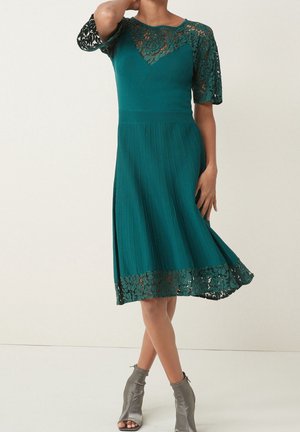 Femme portant une robe tricotée bleu sarcelle avec des manches et un ourlet en dentelle, associée à des bottines argentées à bouts ouverts, debout devant un fond uni.