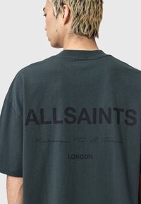 Tamsiai žalia medvilninė marškinėliai, kuriuose užpakalinėje pusėje bold juodu tekstu užrašyta "ALLSAINTS", o po juo – ranka rašyta parašas ir "LONDON".