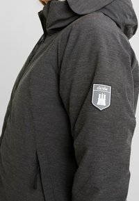 Chaqueta gris texturizada con cierre de cremallera y capucha. Cuenta con un parche con un diseño de castillo en la manga izquierda. Material ligero y transpirable.