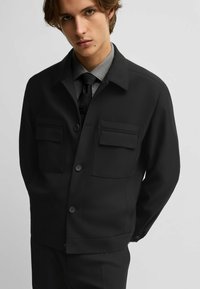 Giacca nera con colletto a camicia, due tasche sul petto e chiusura con bottoni. Tessuto liscio e vestibilità sartoriale, abbinata a una camicia formale grigia.