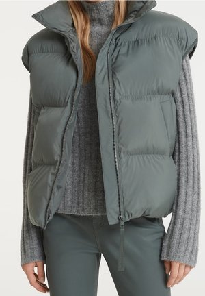 Woman wearing a gray quilted sleeveless puffer vest over a ribbed gray sweater and matching gray pants, standing against a plain background.