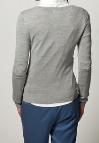 Grauer gestrickter Pullover mit langen Ärmeln, mit einer taillierten Passform und geripptem Saum. Über einem weißen Hemd mit Kragen getragen, kombiniert mit blauen Hosen.