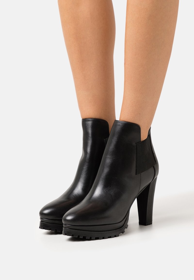 Femme portant des bottines en cuir noir à talons hauts, bouts ronds et panneaux élastiques sur les côtés, debout sur une surface blanche.