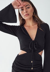 Black dress with long bell sleeves, V-neckline, ruched detailing, and cut-out sides. Features a drawstring with gold accents.