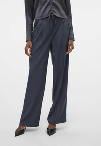Brede broek in donkergrijze stof met een soepele textuur en voorplooien, gestyled met een zwart blouse en puntige hoge hakken.