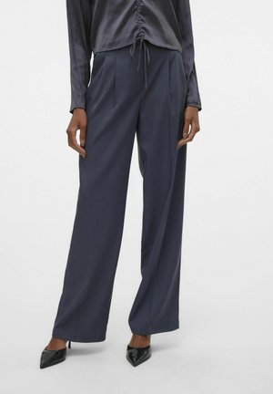 Brede broek in donkergrijze stof met een soepele textuur en voorplooien, gestyled met een zwart blouse en puntige hoge hakken.