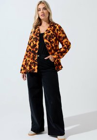 Giacca tie-dye arancione e nera con maniche lunghe e apertura frontale, abbinata a pantaloni neri a vita alta e gamba larga e scarpe di colore chiaro.