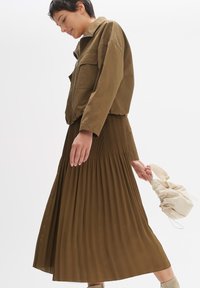 Veste zippée marron avec poches avant, assortie à une jupe longue plissée marron. Sac à cordon de couleur neutre à la main. Textures douces et fluides.