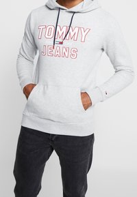 Sudadera con capucha gris con cordón azul marino, con un gran logo rojo de "TOMMY JEANS". Incluye bolsillos tipo canguro y puños acanalados.
