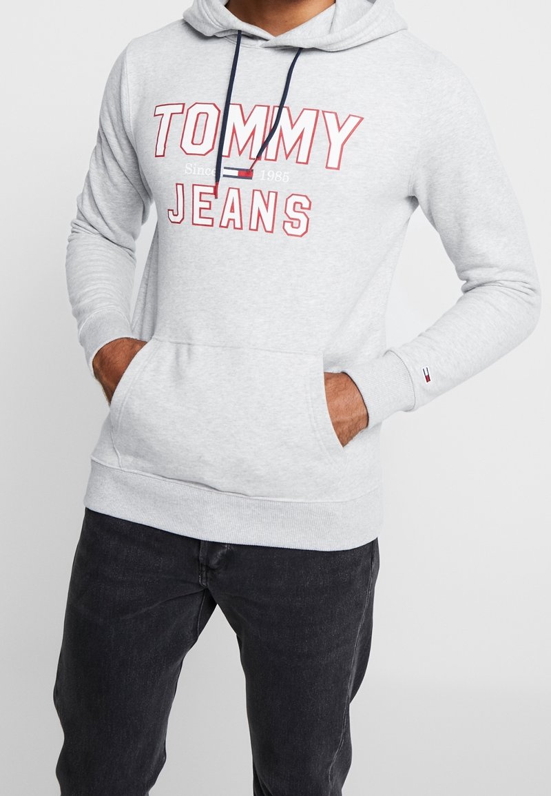 Sudadera con capucha gris con cordón azul marino, con un gran logo rojo de "TOMMY JEANS". Incluye bolsillos tipo canguro y puños acanalados.