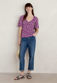 Fioletowa bluzka w kropki z krótkimi falbanowymi rękawami, zestawiona z niebieskimi dżinsami z podwiniętymi mankietami i szarymi sandałami. Trzyma słomkowy kapelusz.